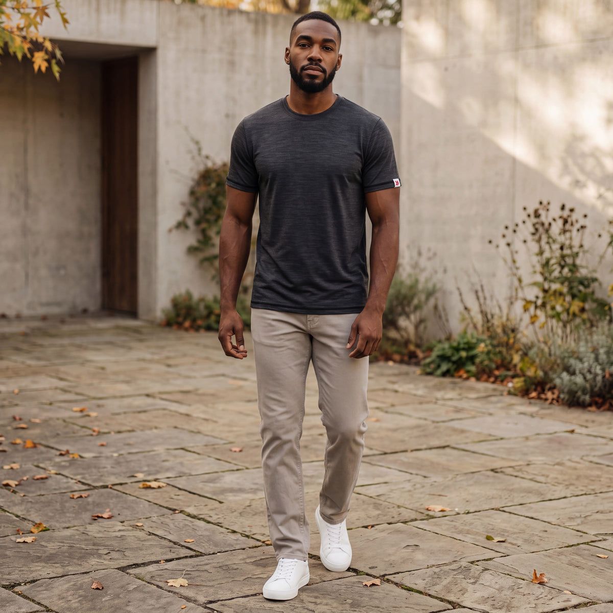 Man walking through a stone courtyard in the Asphalt MOVE Sport Apparel men’s Merino Short Sleeve with stone trousers and white trainers; original sleeve tab retained.