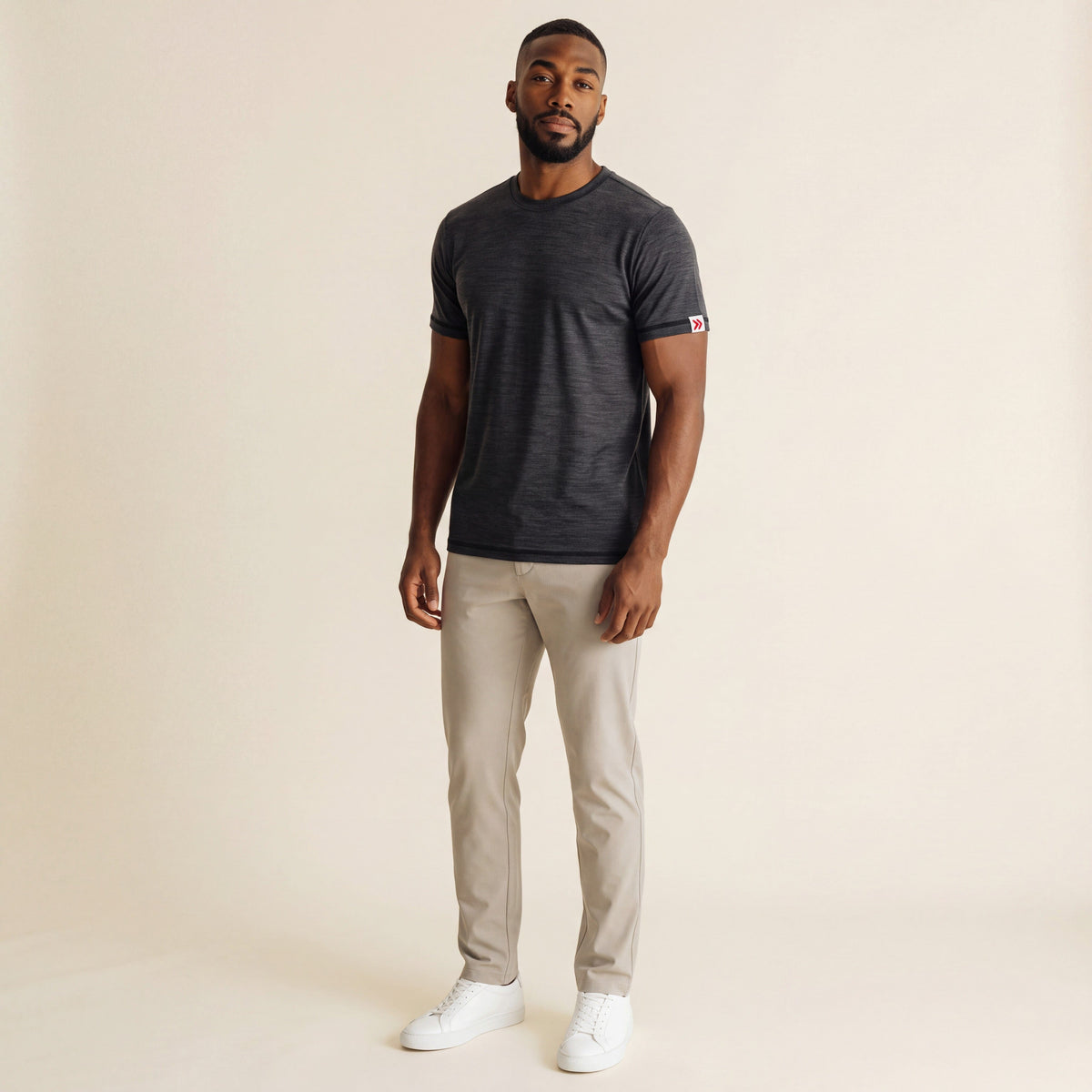 Full-body studio view of a man wearing the Asphalt MOVE Sport Apparel men’s Merino Short Sleeve with stone trousers and white trainers; original red-double-chevron sleeve tab retained.