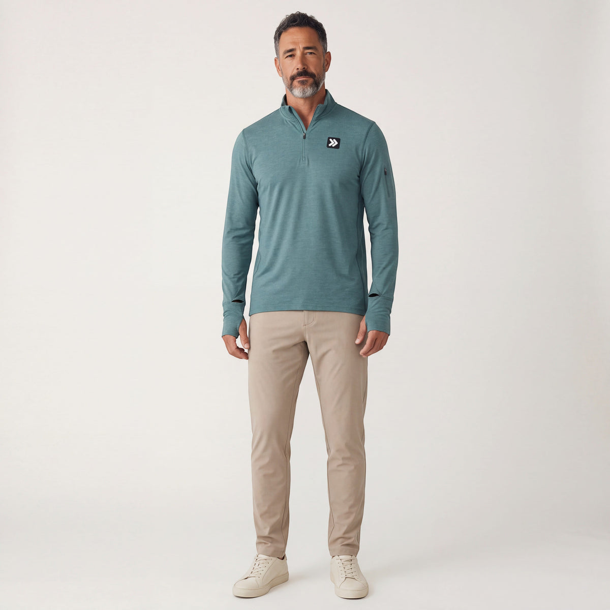 Full-body studio portrait of a male model wearing the Heather North Atlantic MOVE Sport Apparel Essential Quarter Zip Pro, sand trousers and off-white sneakers.