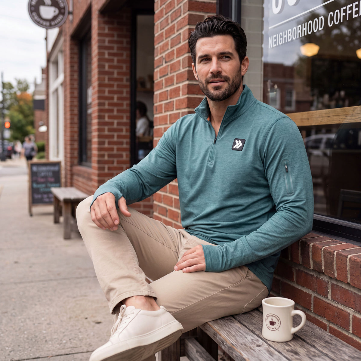 Man wearing the Heather North Atlantic MOVE Sport Apparel Essential Quarter Zip Pro with sand trousers, seated outside a coffee shop.