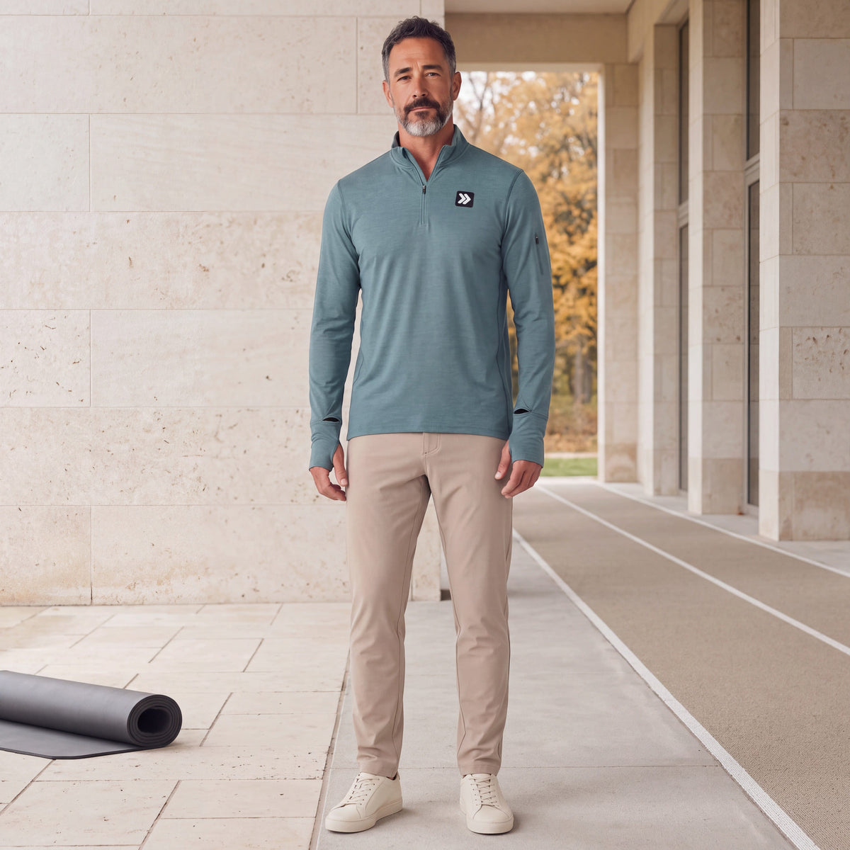 Man wearing the Heather North Atlantic MOVE Sport Apparel Essential Quarter Zip Pro with sand trousers and off-white sneakers at a limestone athletic recovery terrace beside an autumn running track.