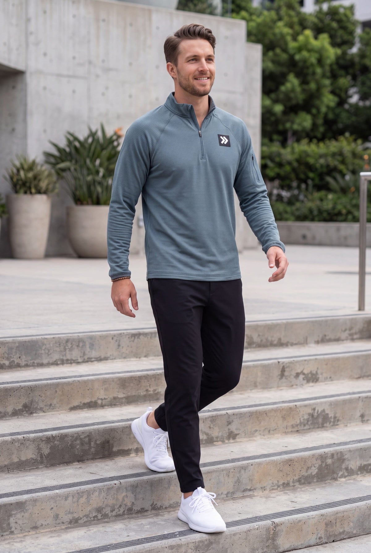 Male model walking on concrete steps wearing MOVE Sport Apparel men’s Balsam Green waffle quarter zip, black training pants and white athletic sneakers.