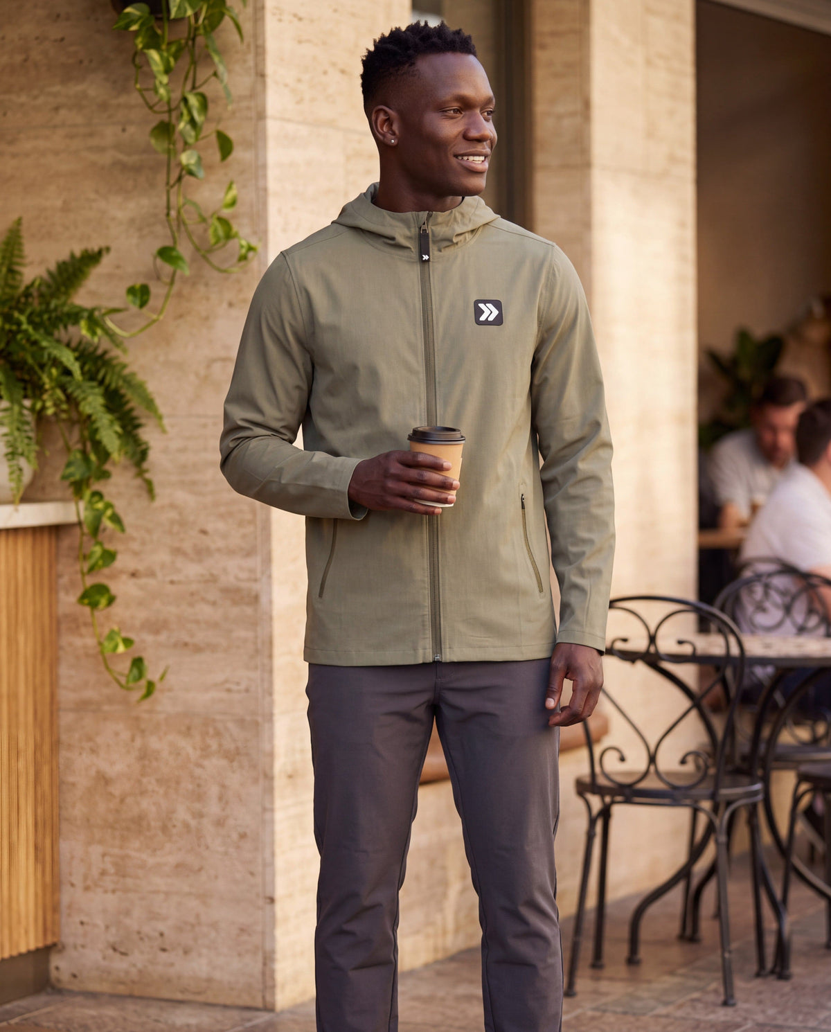 Male model wearing the MOVE Sport Apparel men’s Cactus hooded soft shell jacket outside a stone cafe, holding a coffee.