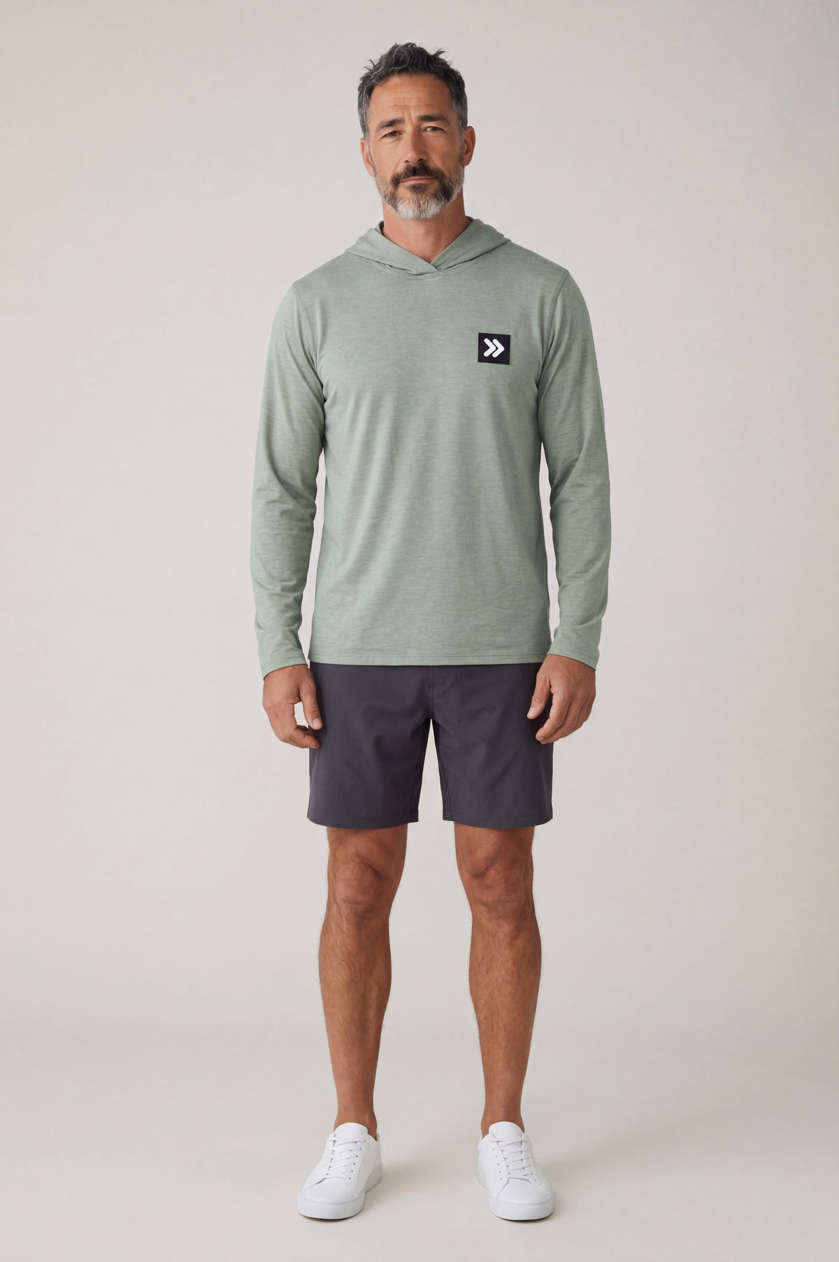 Full-body studio view of a man wearing the Seagrass MOVE Sport Apparel men’s Essential Lightweight Sun Hoodie with charcoal shorts and white trainers; original dark square and white double-chevron chest embellishment retained.