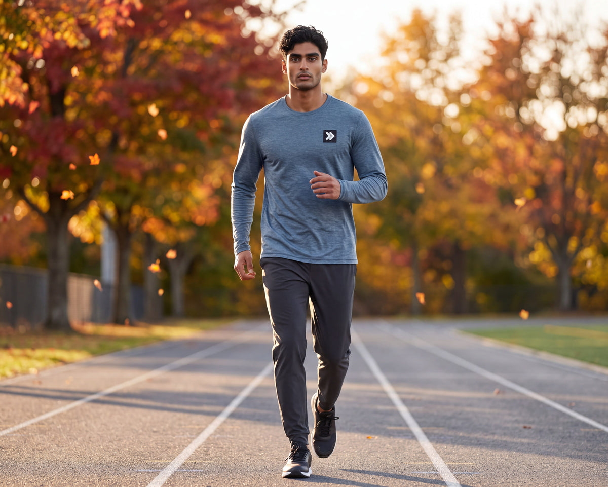 Man warming up on an outdoor track in autumn wearing MOVE Sport Apparel men’s Blue Mirage Black Label long sleeve top and plain charcoal running joggers; original black-and-white chest embellishment retained.