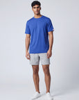Full-body studio view of a man wearing the Surf the Web blue LuxDry Training T-Shirt with light grey shorts and white trainers; original sleeve tab retained.