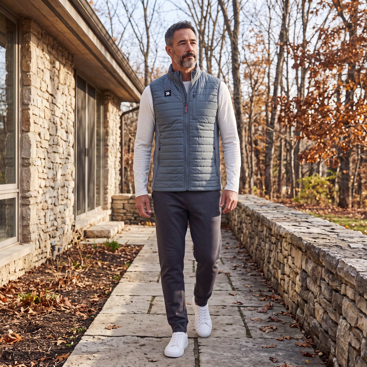 Man wearing the grey MOVE Sport Apparel Men’s down vest over a white long-sleeve top, walking beside stone architecture and autumn trees.