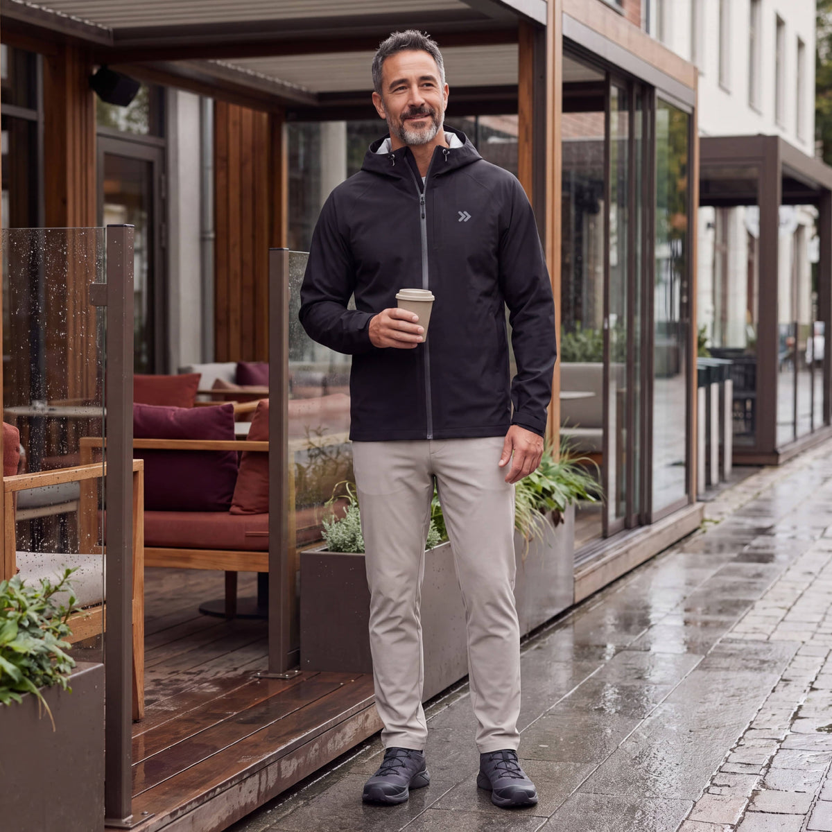 Man wearing the black MOVE Sport Apparel waterproof jacket while holding coffee on a rainy café terrace.