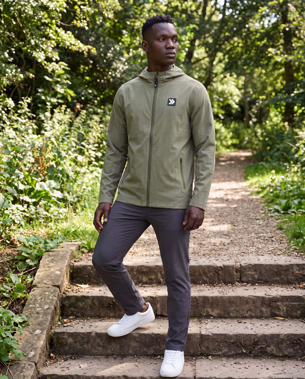 MOVE Sport Apparel men’s Soft Shell Jacket: Male model wearing the Cactus hooded jacket and charcoal trousers on stone steps beside a green park trail.