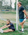 MOVE sport apparel Agility 6 inch training shorts in teal worn by a man stretching by a soccer goal on a field.