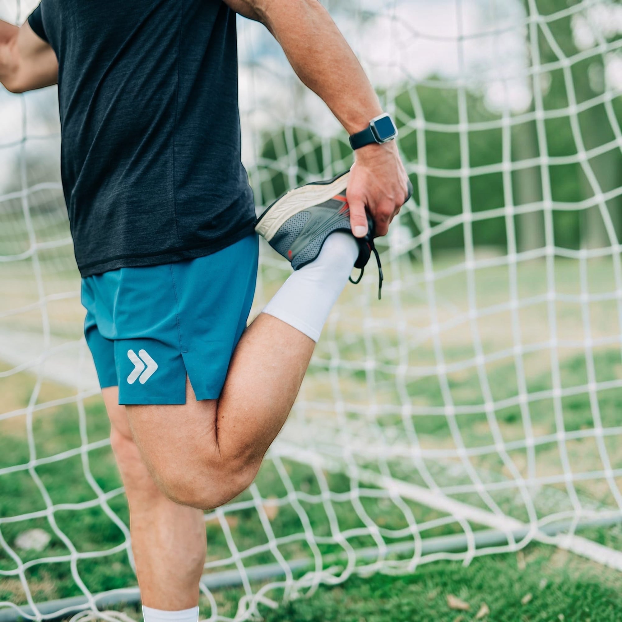 MOVE sport apparel blue 6 inch training shorts with white logo detail worn by a man stretching on a soccer field