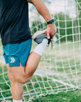 MOVE sport apparel blue 6 inch training shorts with white logo detail worn by a man stretching on a soccer field