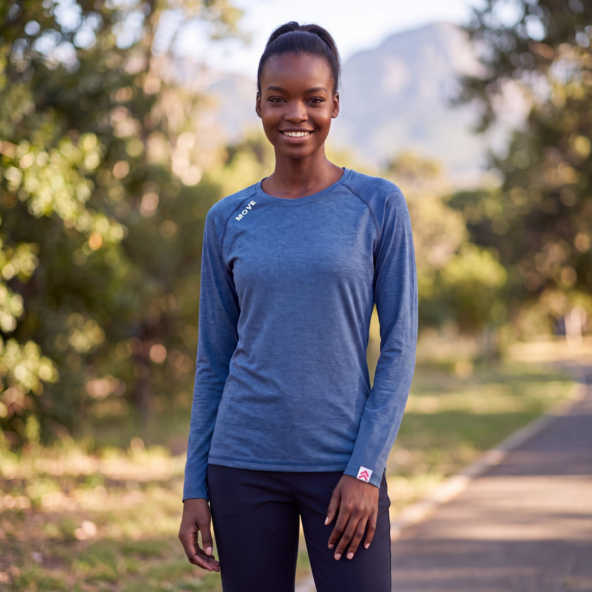 A woman wearing a MOVE sport apparel blue long sleeve shirt with a crew neck, standing outdoors in a park.