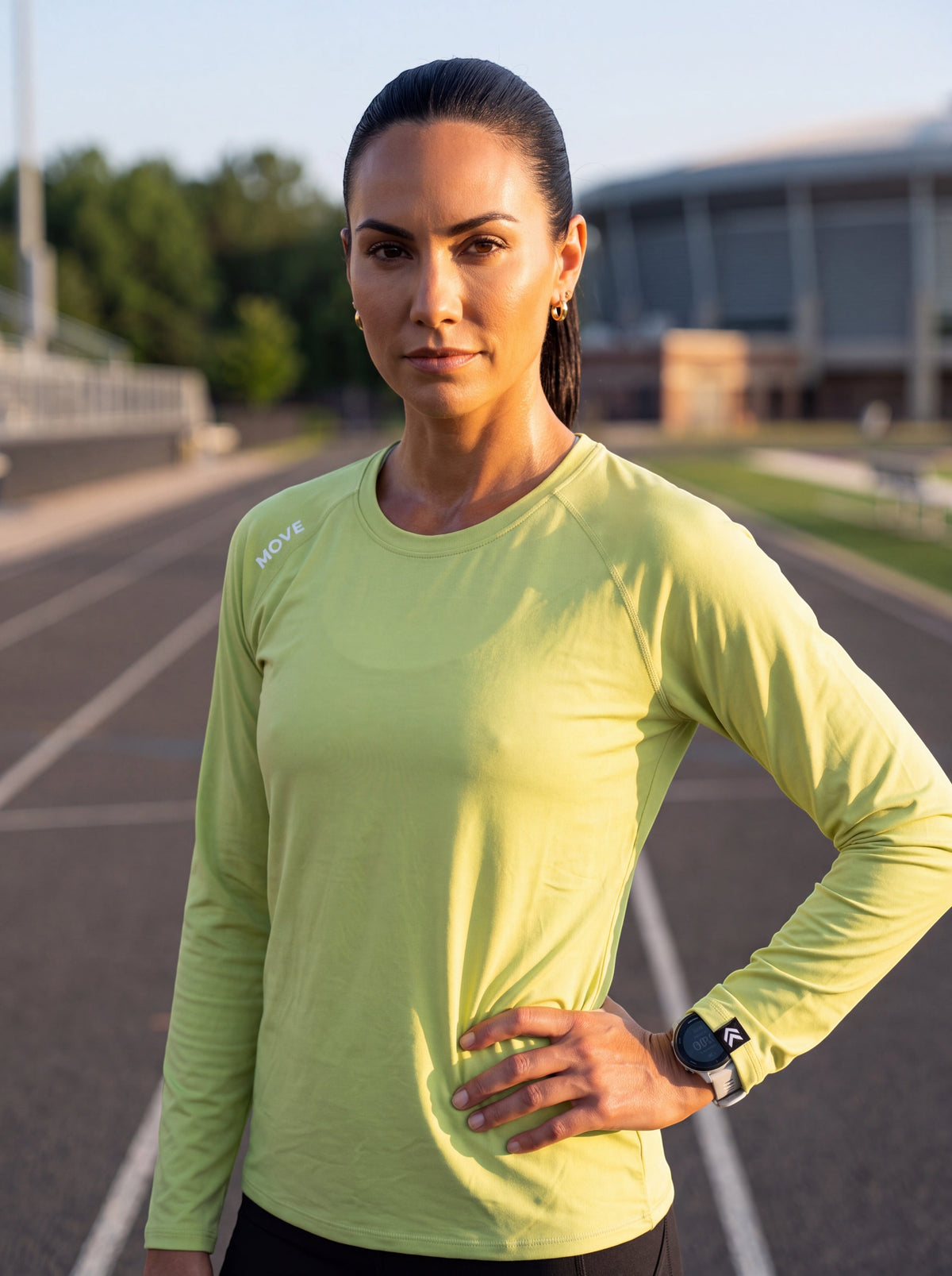A woman wearing a lime green MOVE sport apparel long sleeve shirt on a running track at sunset.