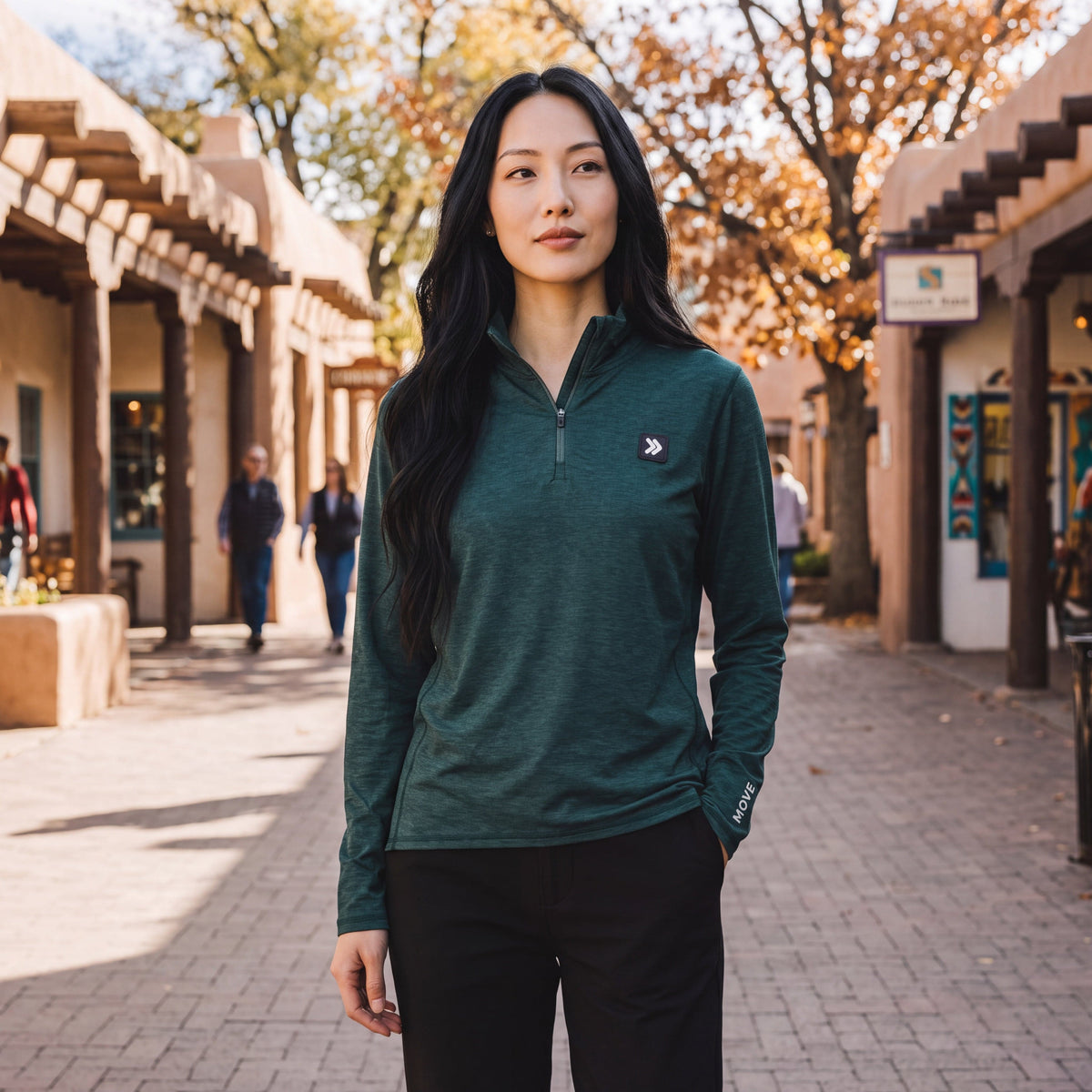 A woman wearing a MOVE sport apparel dark green quarter zip pullover with reflective logos on a sunny outdoor street.