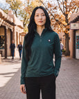 A woman wearing a MOVE sport apparel dark green quarter zip pullover with reflective logos on a sunny outdoor street.