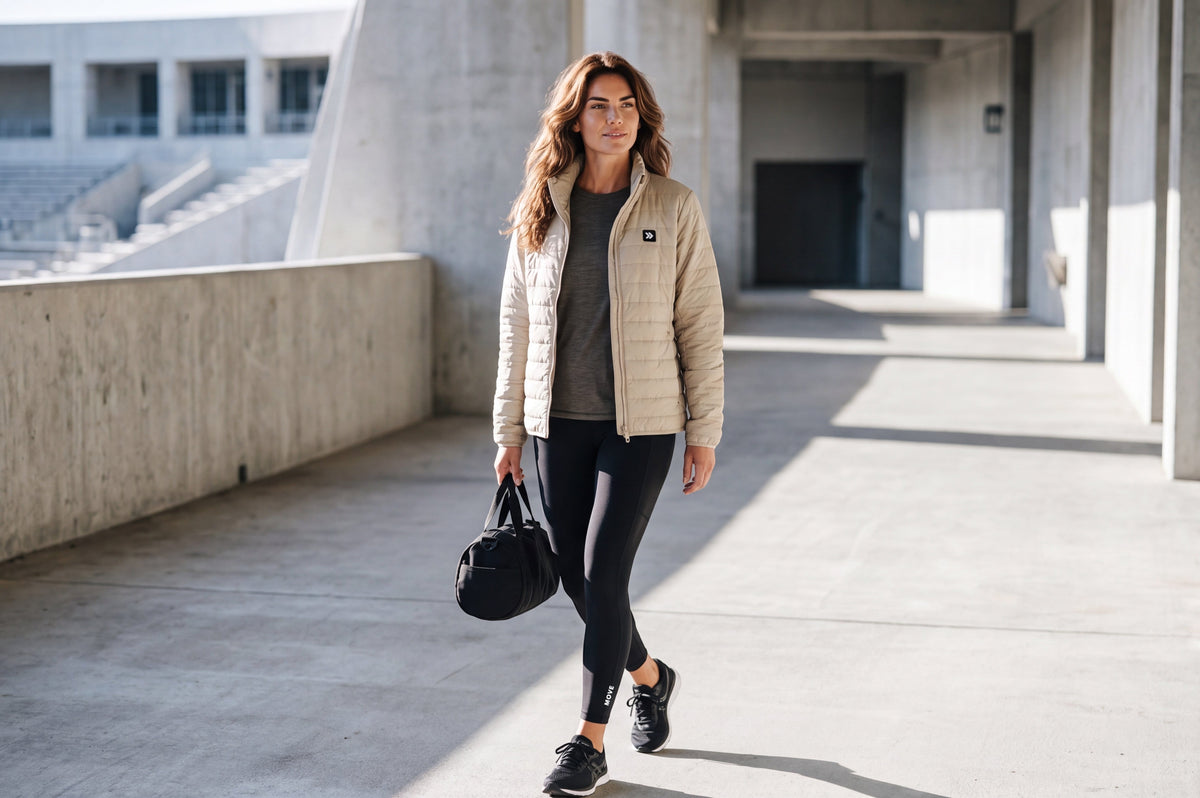 Woman wearing a MOVE sport apparel beige lightweight quilted down jacket with a high neck, leggings, and gym bag.