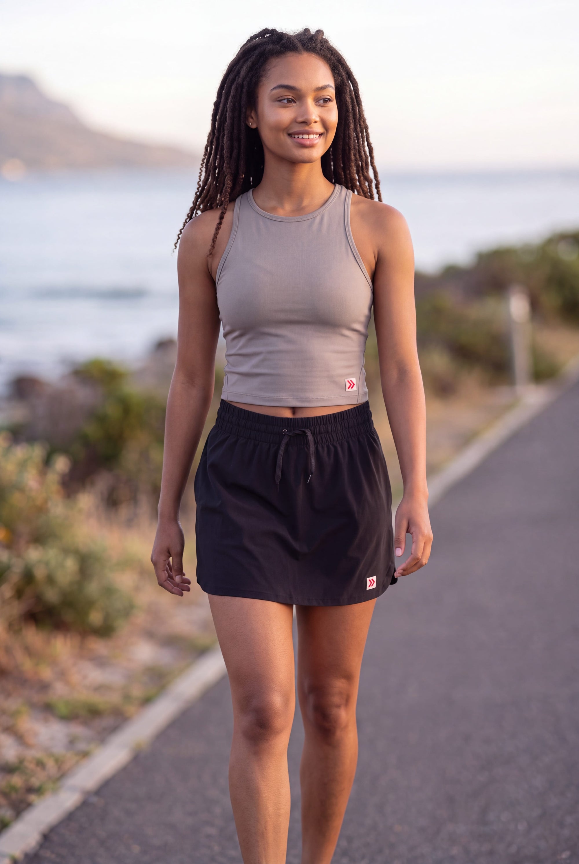A woman wearing a MOVE sport apparel grey cropped tank top and black athletic skirt while walking outdoors.