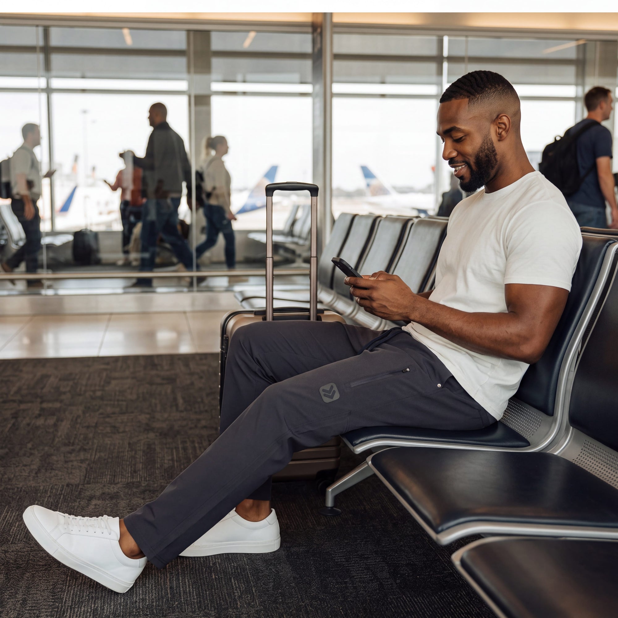 A man wearing MOVE sport apparel charcoal grey ripstop pants sitting in an airport terminal using his smartphone.