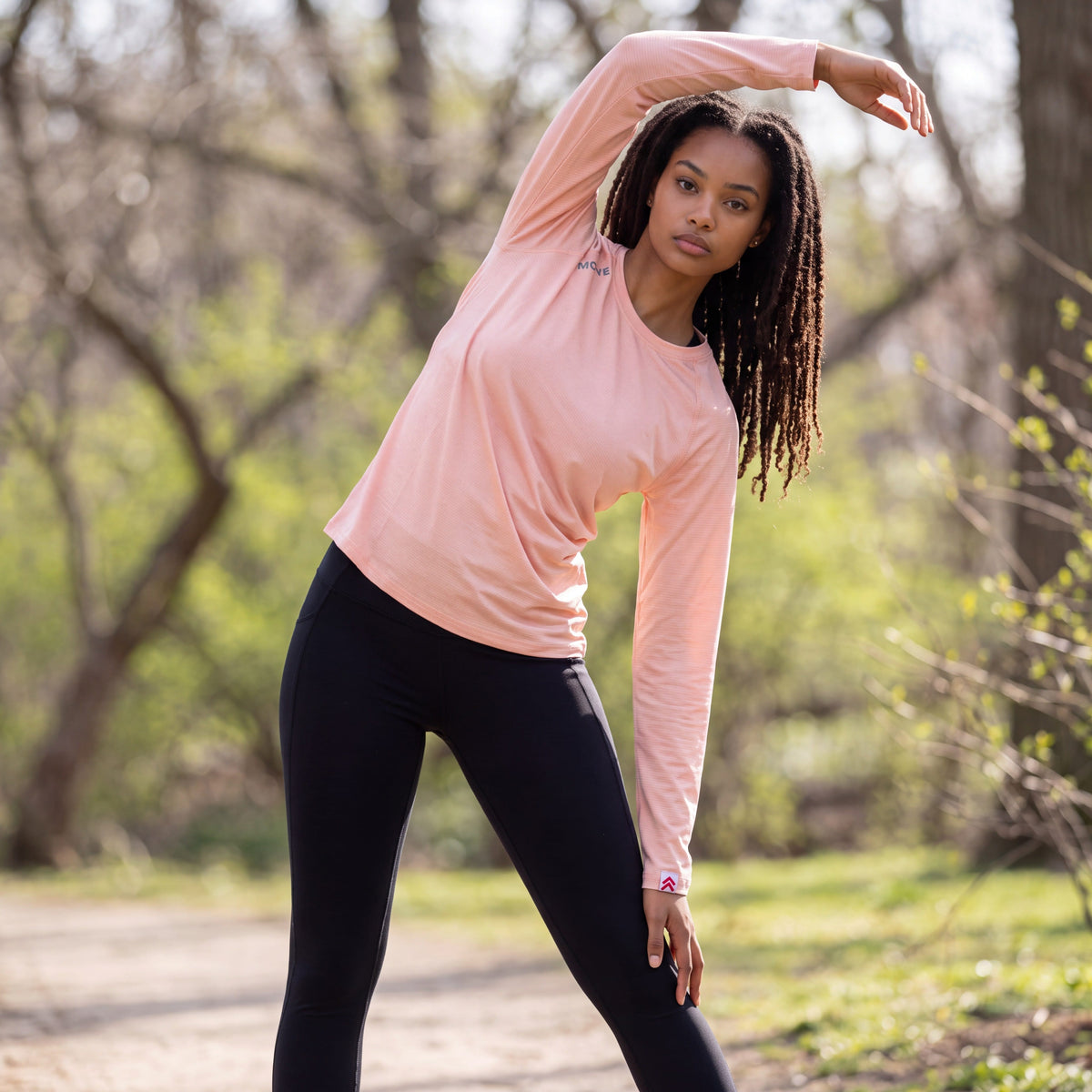 Woman performing a standing side stretch in a park while wearing the MOVE Sport Apparel Long Sleeve LuxDry Performance Crew Neck Shirt in Creole Pink.