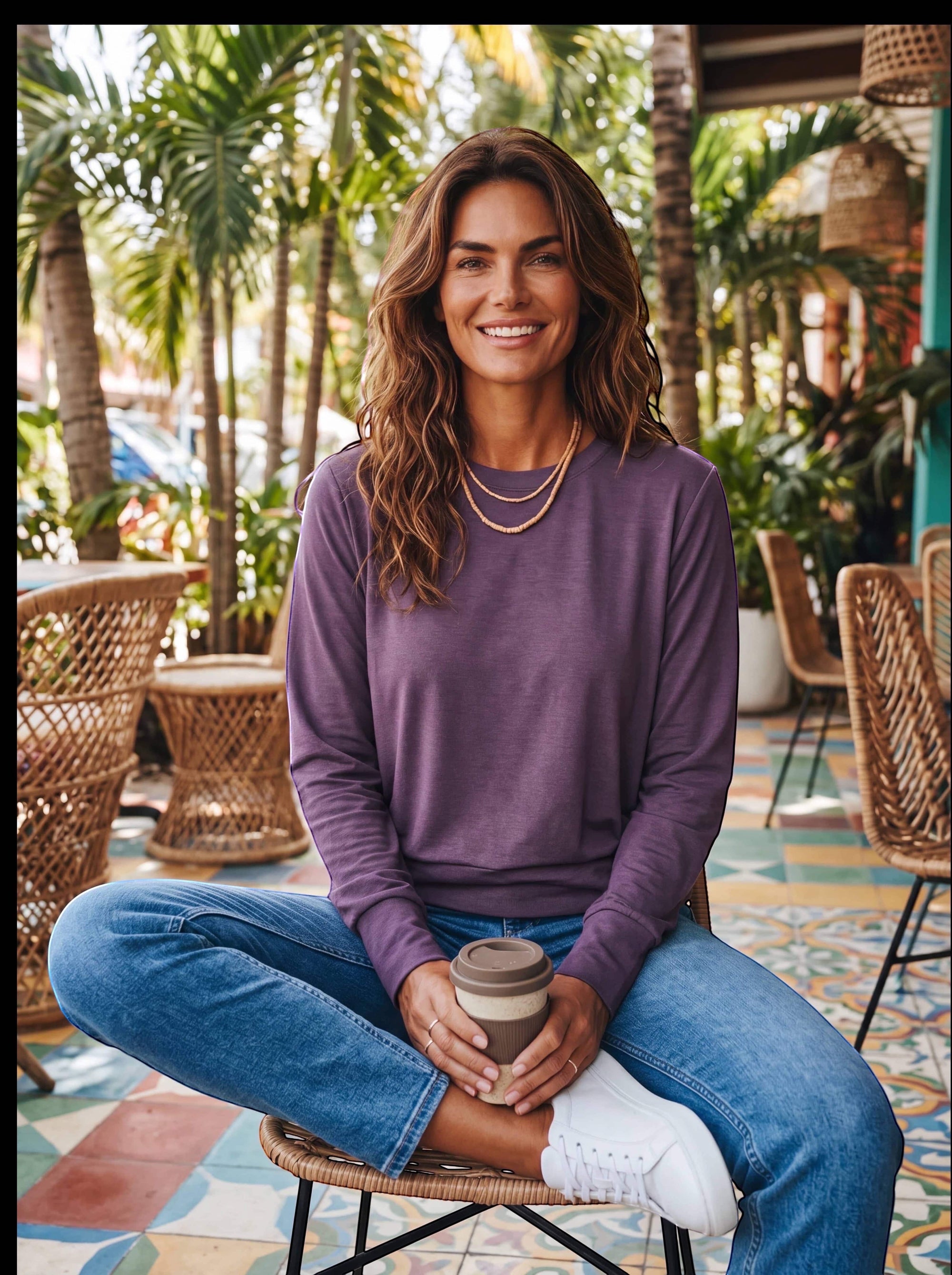 A woman wearing a MOVE sport apparel purple long sleeve crewneck pullover with blue jeans in a tropical cafe setting.