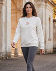Woman wearing the cream MOVE Sport Apparel Logo Sweatshirt with charcoal leggings beside stone architecture and autumn trees.