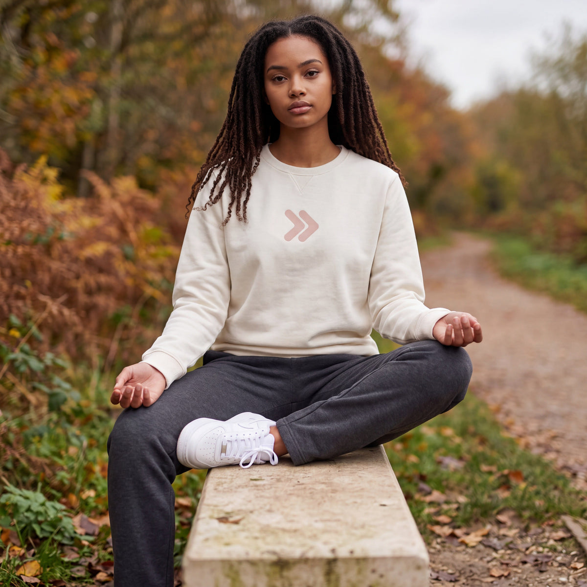 Woman wearing the cream MOVE Sport Apparel Logo Sweatshirt with its blush-pink chest embellishment, seated on a stone bench beside an autumn trail.
