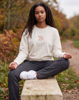 Woman wearing the cream MOVE Sport Apparel Logo Sweatshirt with its blush-pink chest embellishment, seated on a stone bench beside an autumn trail.