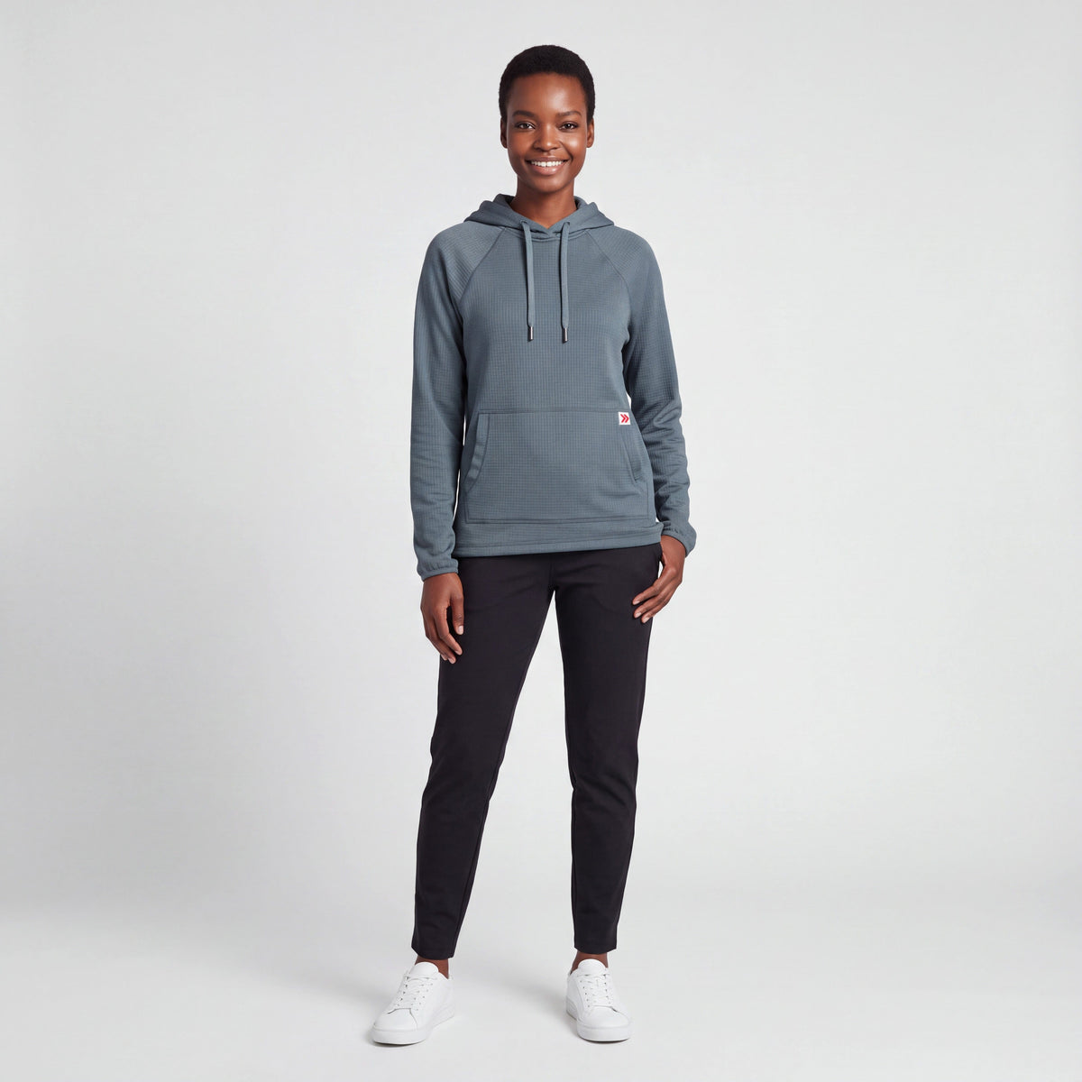 Full-body female studio model wearing MOVE Sport Apparel women’s Balsam Green waffle hoodie with black training pants and white sneakers.