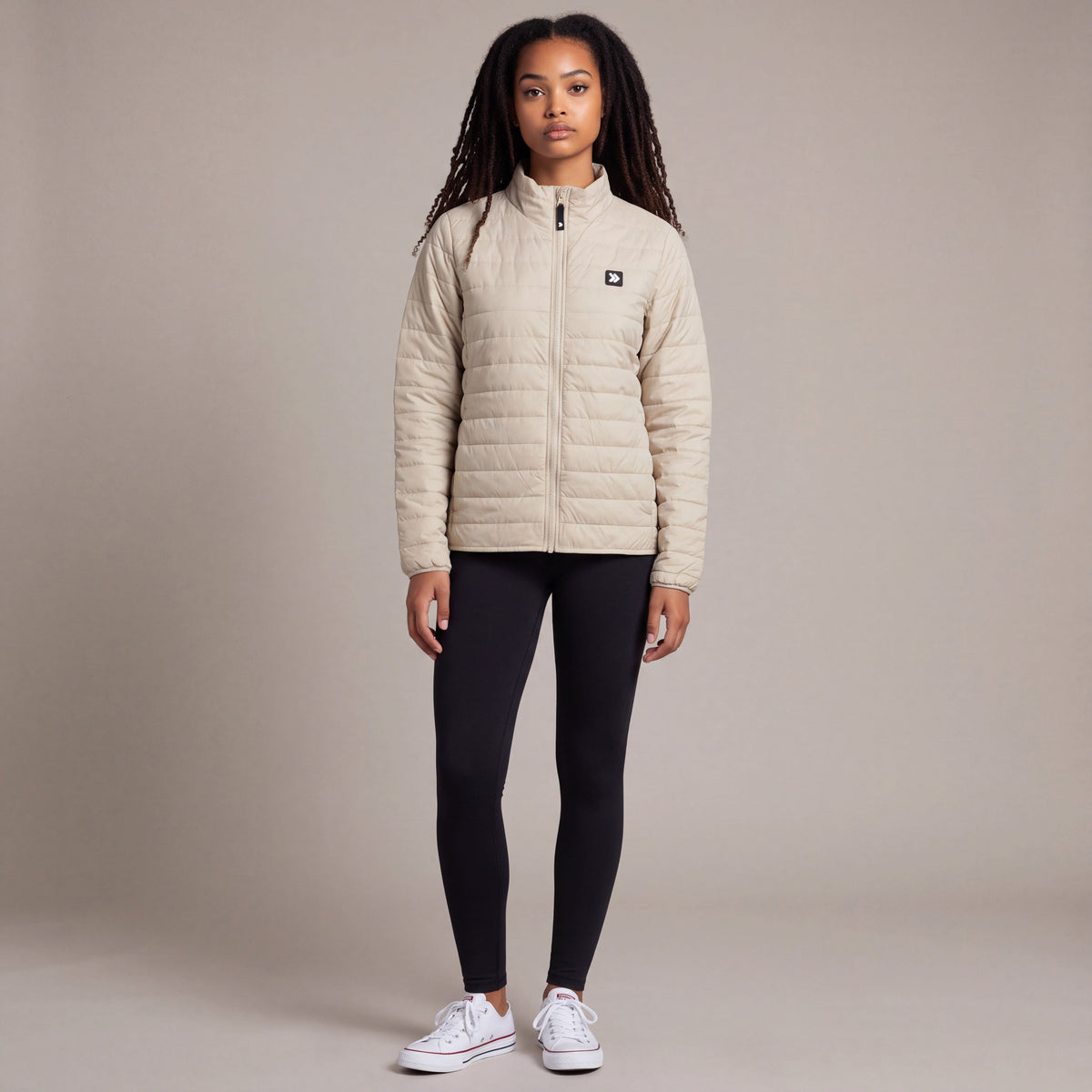Full-body studio view of a woman wearing the Silver Birch MOVE Sport Apparel Women’s Lightweight Down Jacket with black leggings and white trainers, showing the quilted panels and original chest patch.