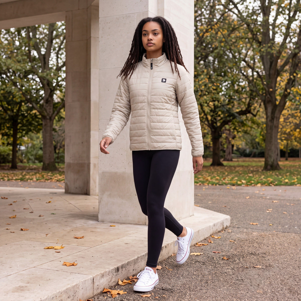 Woman walking beside a limestone park pavilion in autumn, wearing the Silver Birch MOVE Sport Apparel Women’s Lightweight Down Jacket with black leggings and white trainers.