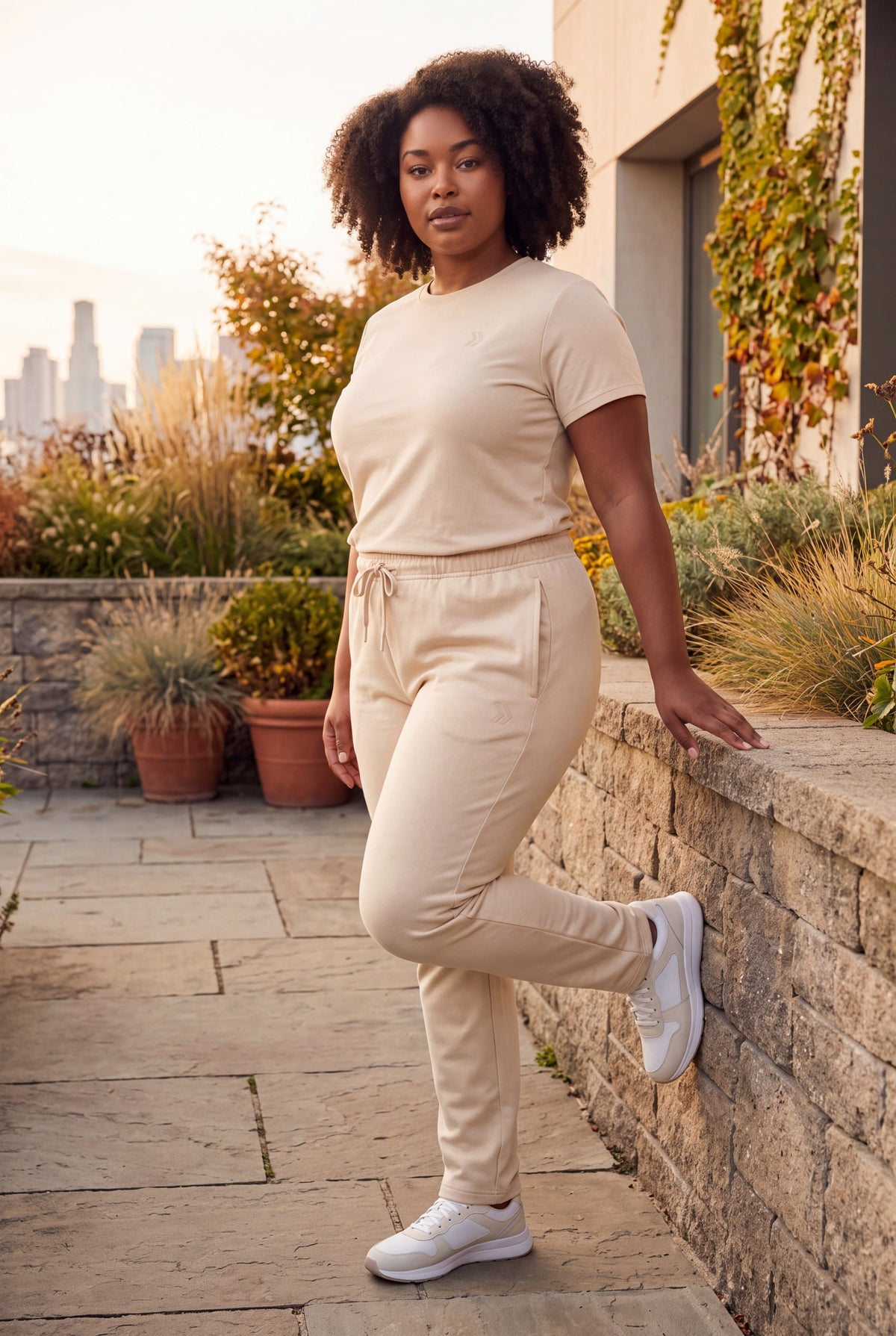Woman wearing MOVE Sport Apparel French Terry Joggers in Natural during a gentle standing stretch beside a stone wall on an autumn terrace.