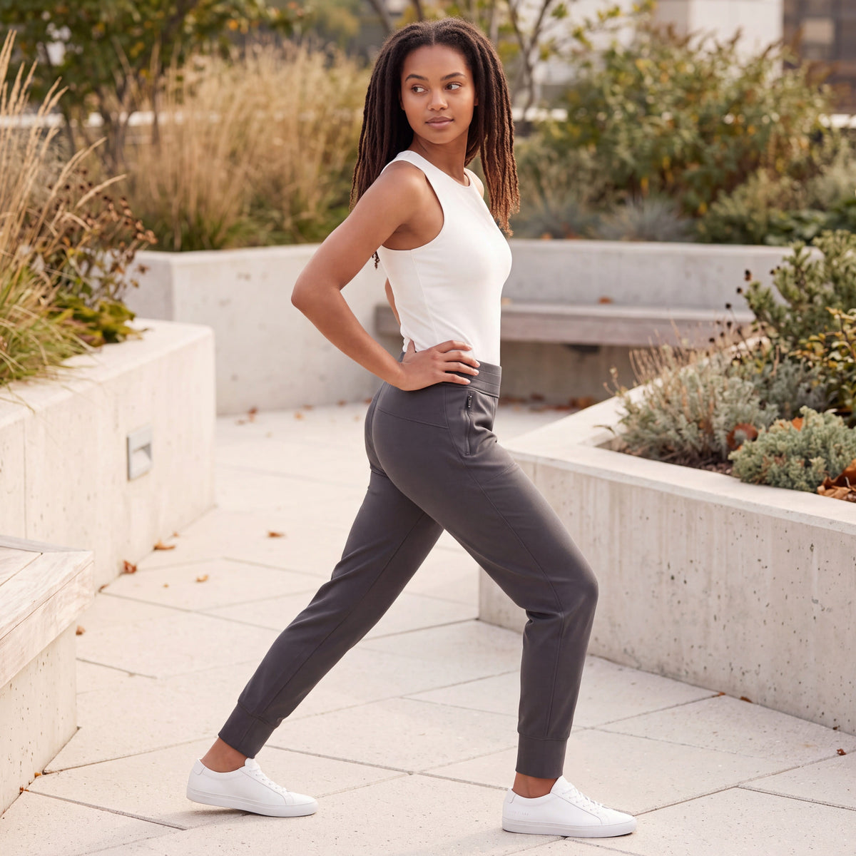 Woman gently stretching outdoors in MOVE Sport Apparel Women’s Cloud Joggers in Soft Black with an ivory sleeveless top on a landscaped terrace in early autumn.