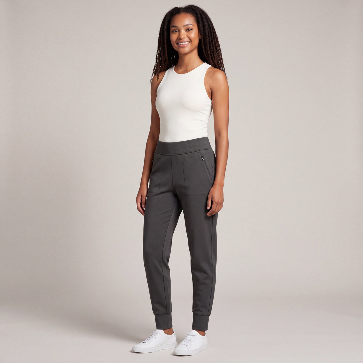 Full-body studio view of a woman wearing MOVE Sport Apparel Women’s Cloud Joggers in Soft Black with an ivory sleeveless top and white trainers, showing the zip pockets and ankle cuffs.