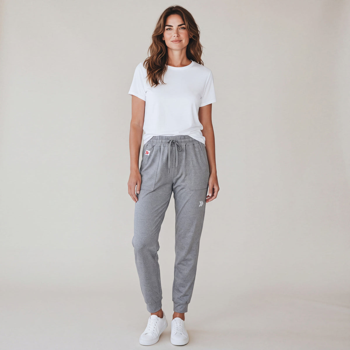 Full-body studio view of a woman wearing grey MOVE Sport Apparel Women’s Essential Joggers with a white tee and trainers, showing the pocket tab and silver reflective thigh embellishment.