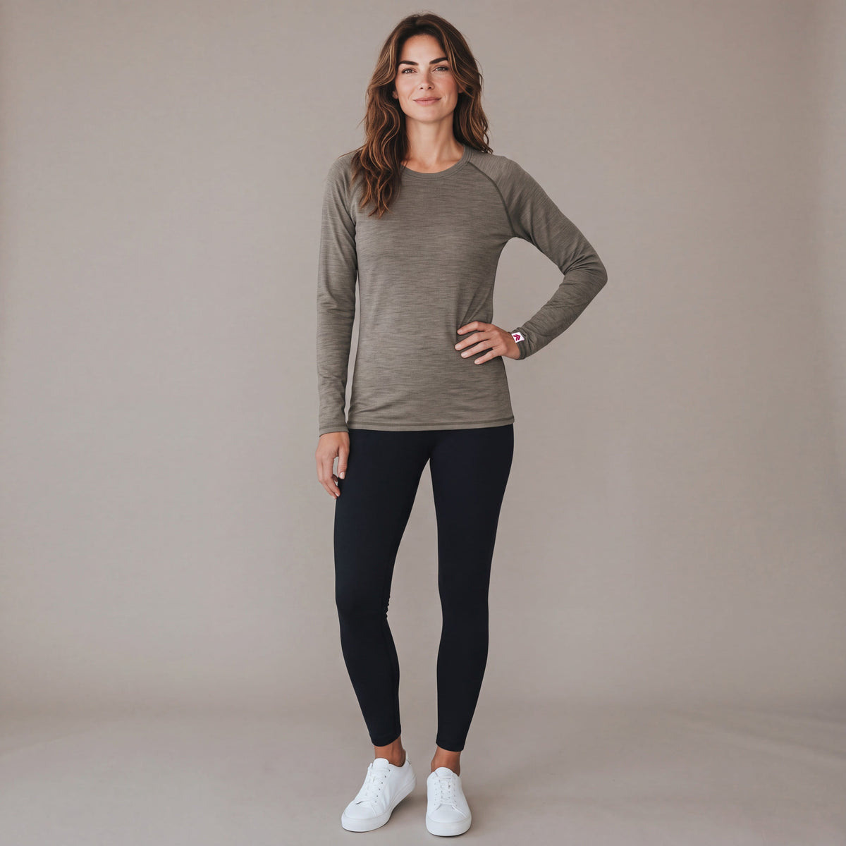 Full body studio view of a woman wearing the MOVE Sport Apparel Women’s Long Sleeve Merino Base Layer Shirt in Smokey Olive with black leggings.