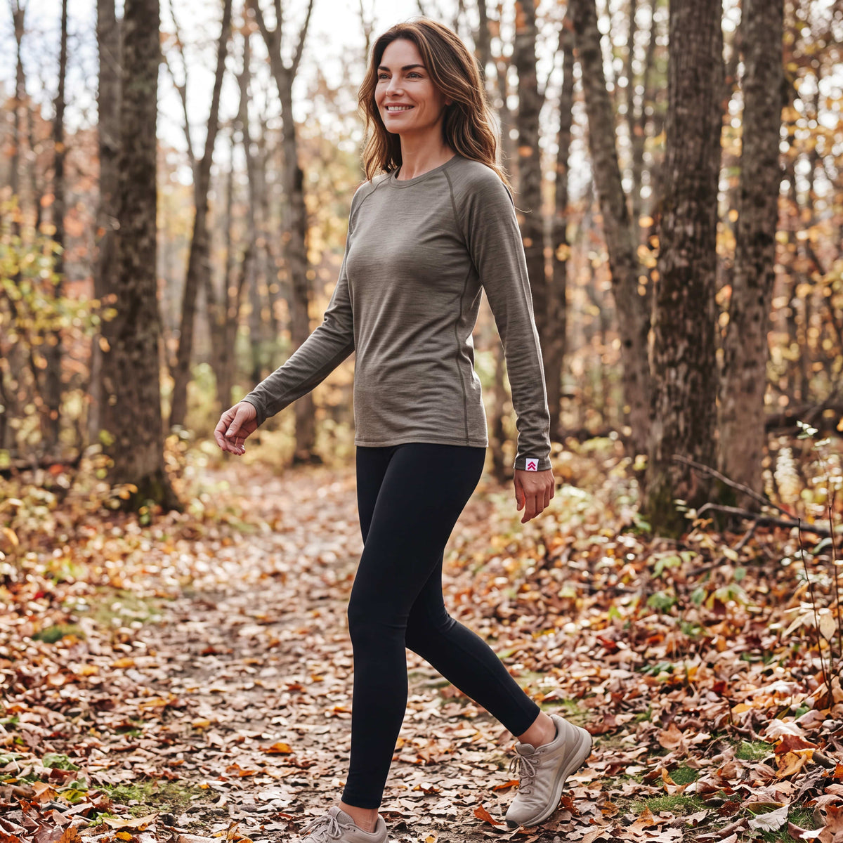 Woman walking along an autumn woodland trail in the MOVE Sport Apparel Women’s Long Sleeve Merino Base Layer Shirt in Smokey Olive, with the original cuff label.