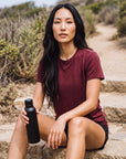 MOVE Sport Apparel women’s Merino Short Sleeve T-Shirt: Woman wearing Oxblood Red Merino Short Sleeve T-Shirt while resting on a stone coastal trail.