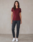 MOVE Sport Apparel women’s Merino Short Sleeve T-Shirt: Full-body studio view of a woman wearing Oxblood Red Merino Short Sleeve T-Shirt with charcoal trousers and white trainers.