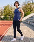 Woman wearing the MOVE Sport Apparel Pure Stretch Full Length Tank Top in Maritime Blue with black leggings beside an outdoor running track in spring.