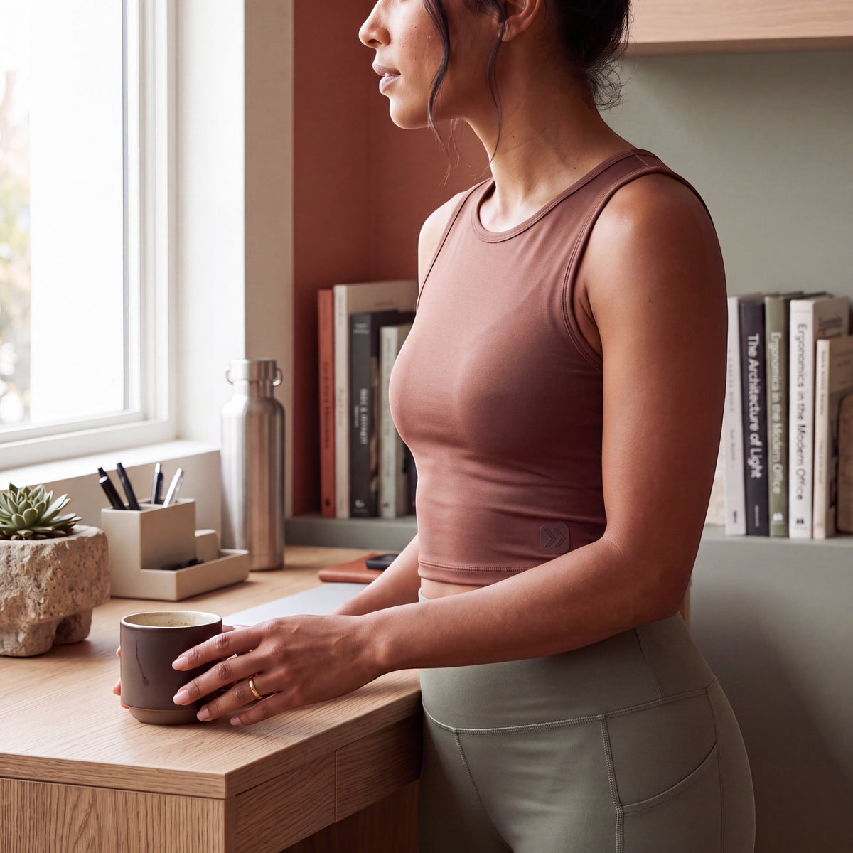 Woman wearing a muted brown cropped tank beside a desk in a softly lit interior.