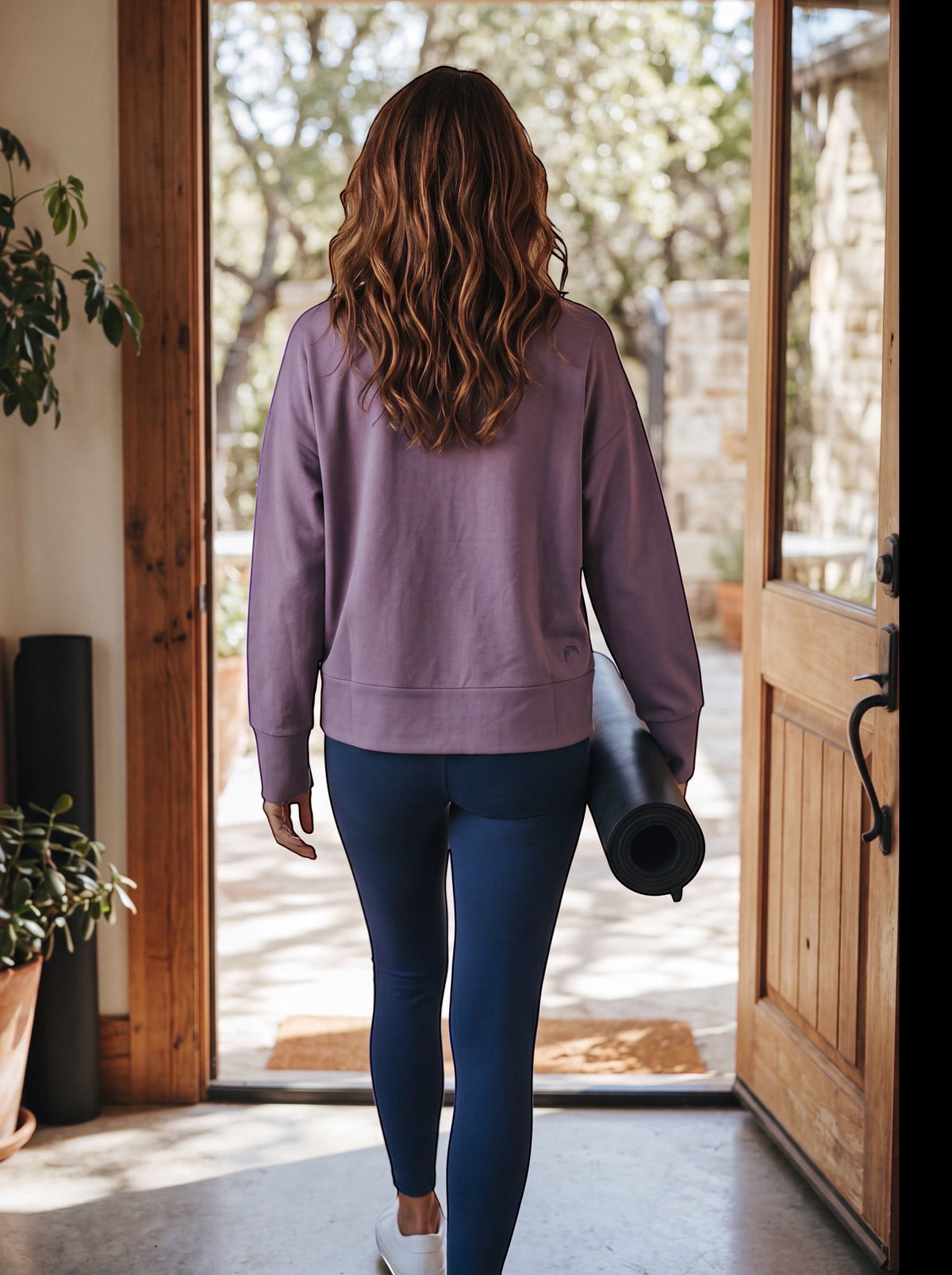 Back view of a woman wearing the MOVE Sport Apparel Women’s Long Sleeve Crewneck Pullover in Gothic Grape while carrying a yoga mat.