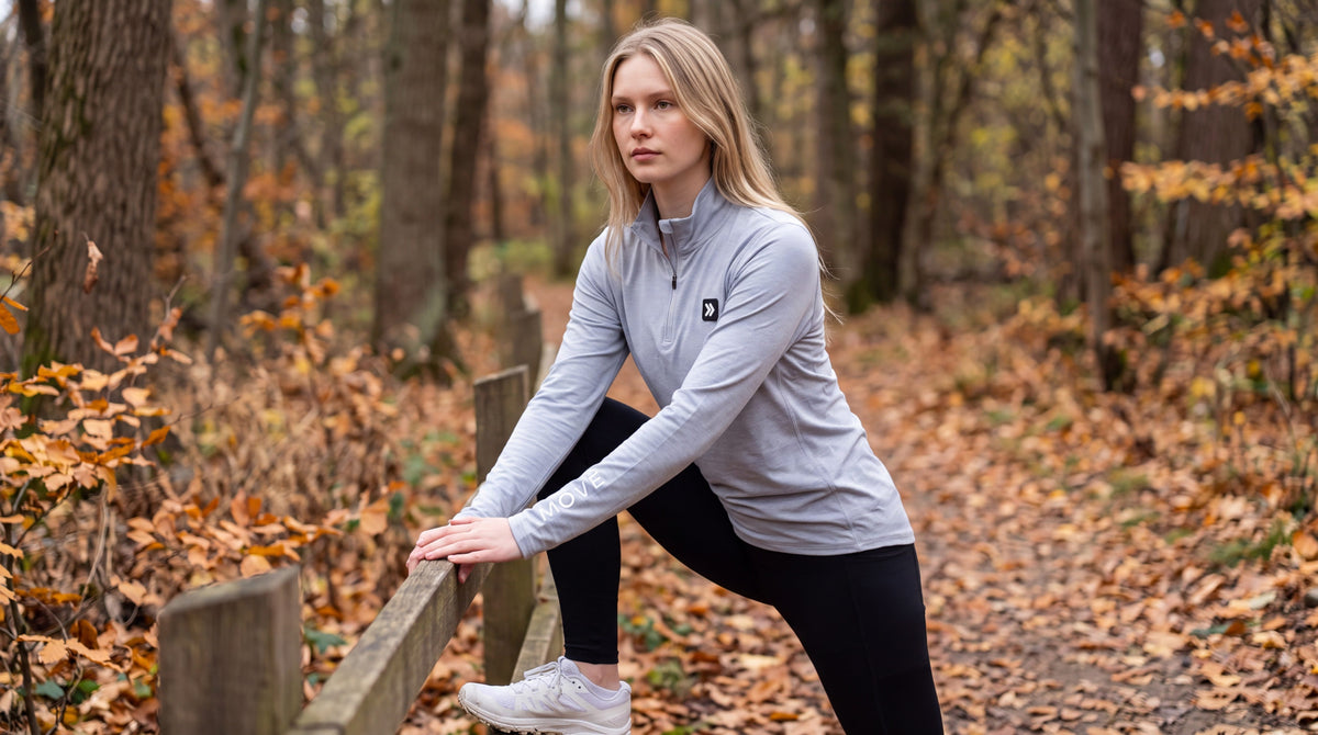 Woman stretching beside a wooden rail on an autumn trail in the MOVE Sport Apparel Women’s Essential Quarter Zip in Silver Cloud, showing the original chest patch and sleeve lettering.