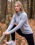 Woman stretching beside a wooden rail on an autumn trail in the MOVE Sport Apparel Women’s Essential Quarter Zip in Silver Cloud, showing the original chest patch and sleeve lettering.