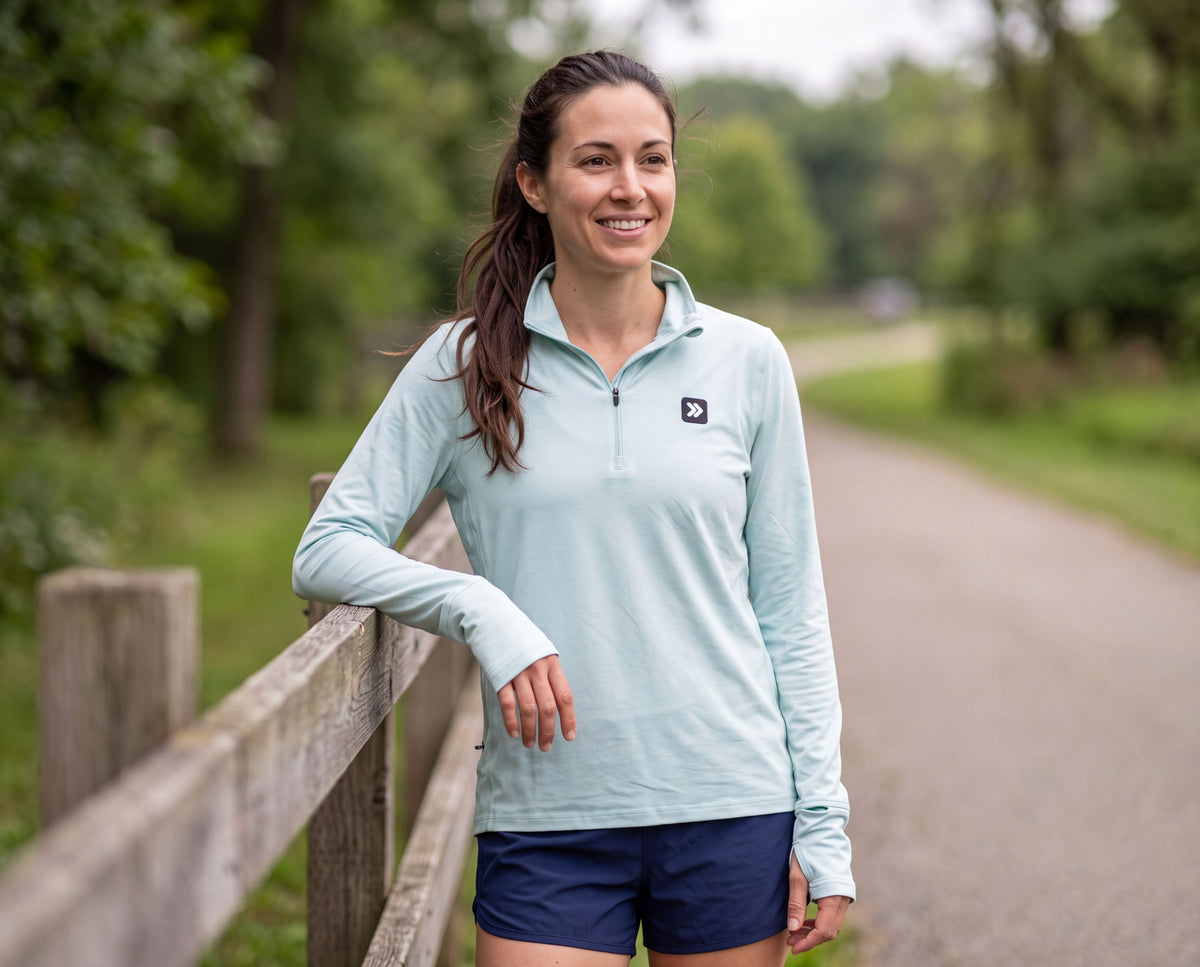 Woman wearing the Heather Skylight MOVE Sport Apparel Essential Quarter Zip Pro with navy running shorts, resting by a wooden rail on a leafy park path.