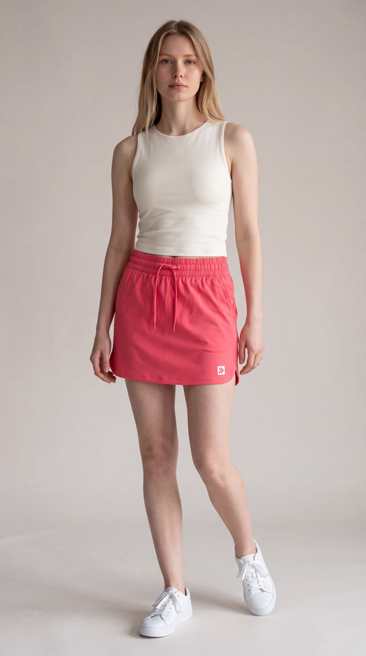 Full-body studio view of a woman wearing MOVE Sport Apparel women’s Skort in Paradise Pink with an ivory sleeveless top and white trainers; original waistband, curved hem and small hem tab retained.