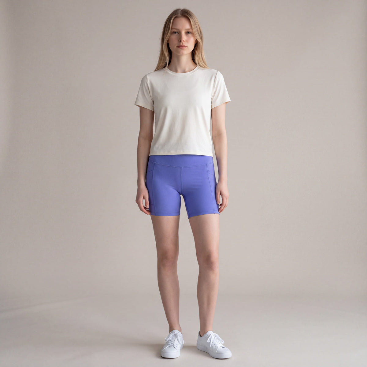 MOVE Sport Apparel Women’s High Rise Athletic Performance Shorts in Deep Periwinkle, front view worn by an adult woman in a neutral studio with a plain ivory T-shirt and white trainers.
