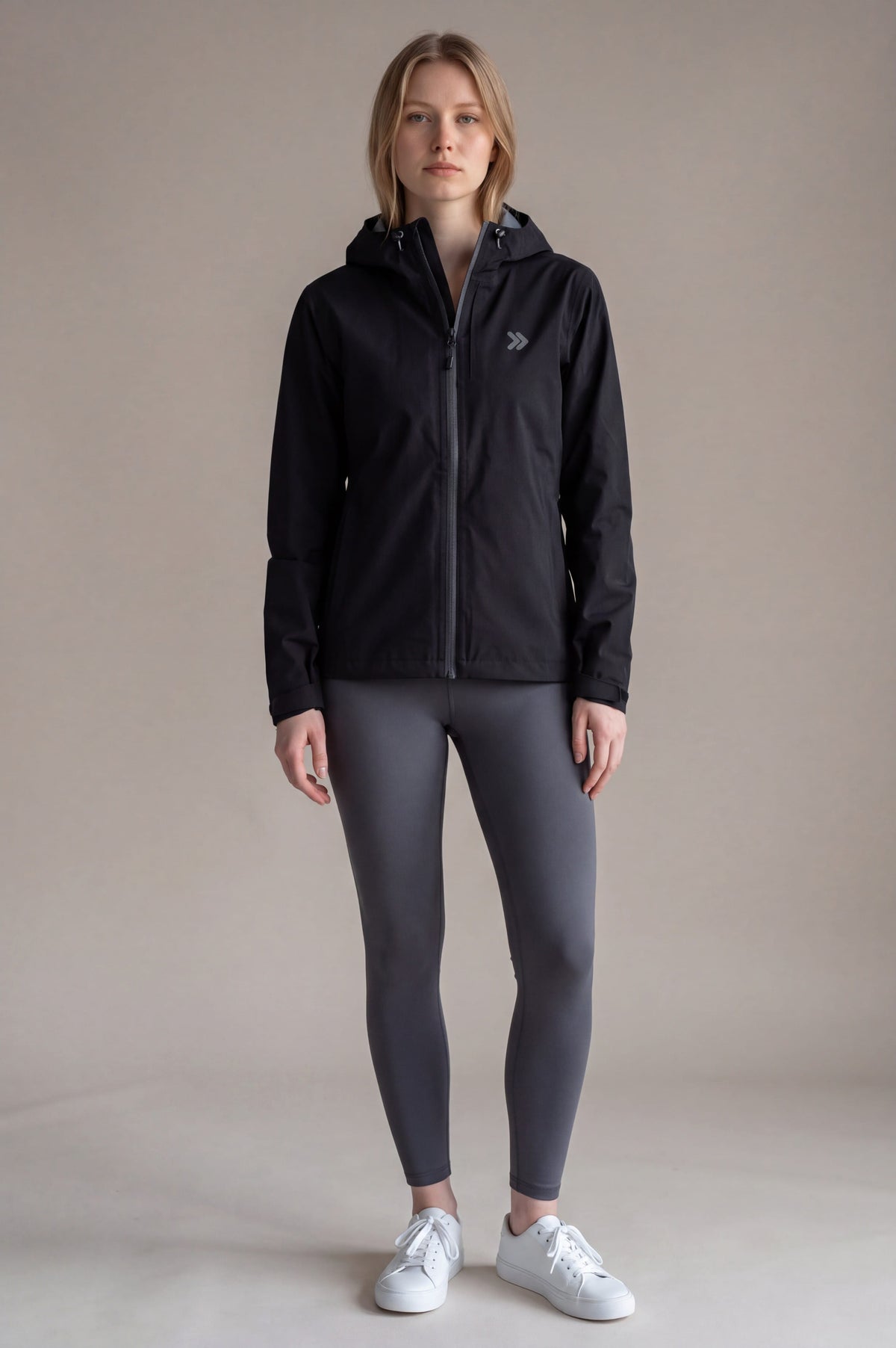 Full-length studio view of a woman wearing the black MOVE Sport Apparel hooded waterproof jacket with charcoal leggings.