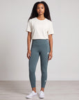 Full-body studio view of a woman wearing MOVE Sport Apparel women’s 25-Inch Running Tights in Balsam Green with an ivory top and white trainers.