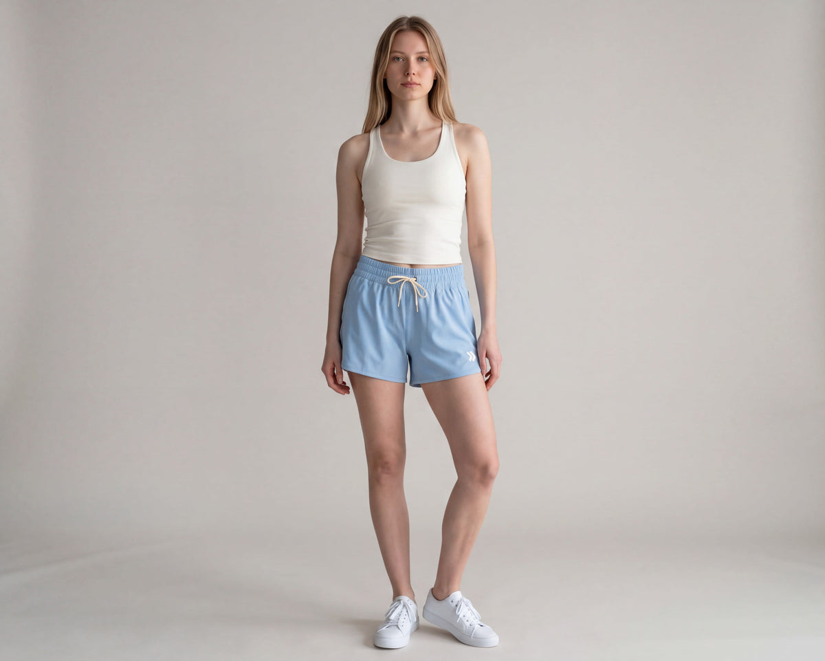 Full-body studio view of a woman wearing Mountain Bluebird MOVE Sport Apparel women’s 4-Inch Running Shorts with an ivory tank and white trainers; original white double-chevron embellishment retained.